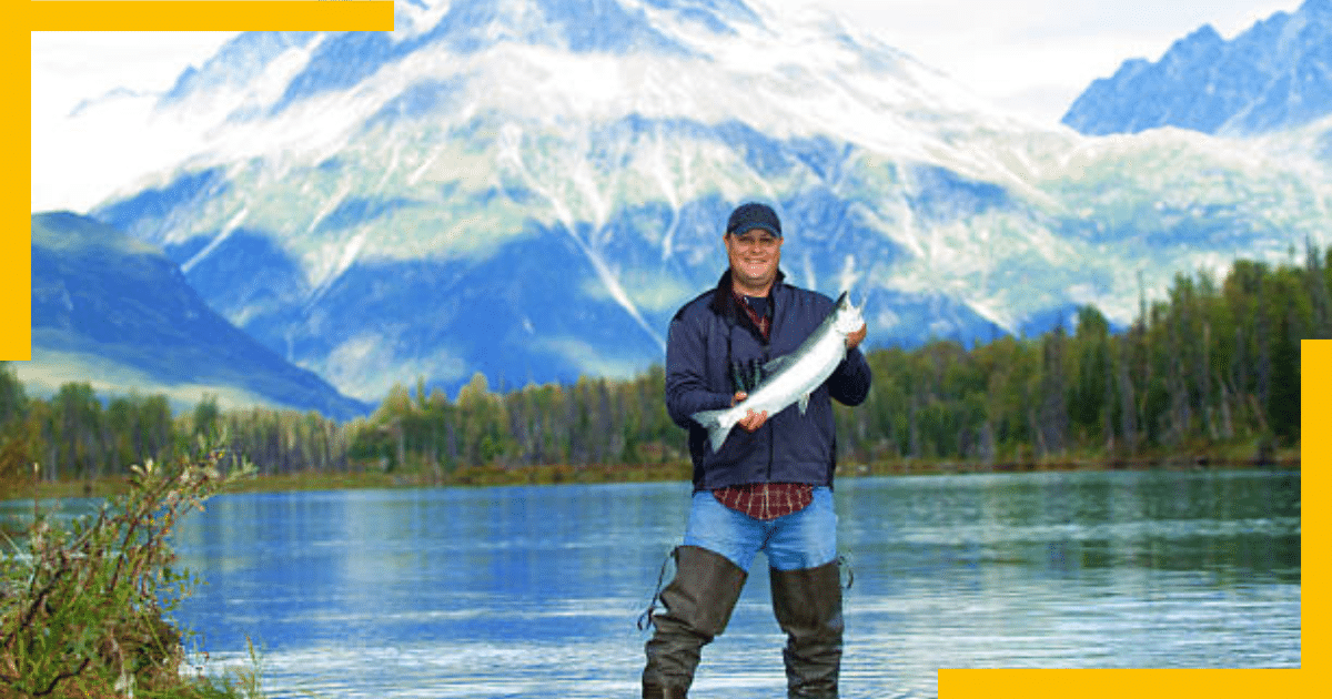 A man holding a fish and standing in front of a lake in Alaska